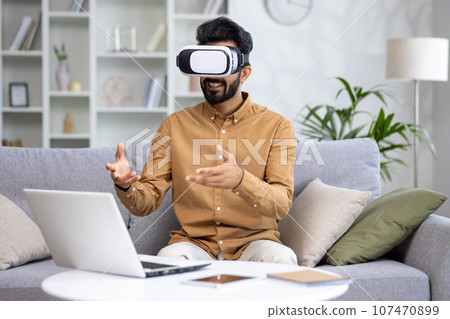 Indian young man sitting at home on sofa in front of laptop, working, studying and talking through virtual mask online. Indian young man sitting at home on sofa in front of laptop, working, studying and talking through virtual mask online. 107470899