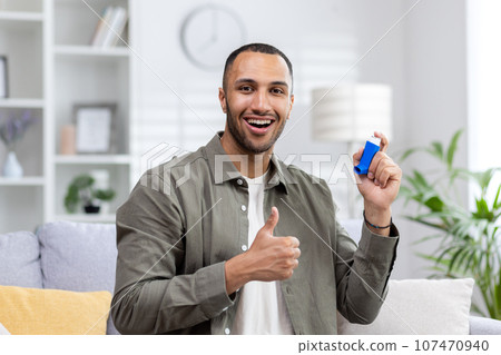 Portrait of a young African American man sitting on the sofa at home, holding an asthma spray in his hand, smiling and pointing super finger at the camera. 107470940