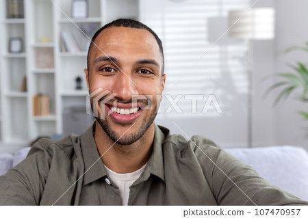 Young African American man standing at home, talking on a video call, recording, smiling at the camera. Close-up photo. Young African American man standing at home, talking on a video call, recording, smiling at the camera. Close-up photo. 107470957