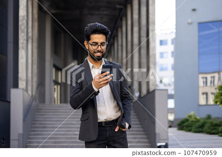 A young businessman in a business suit and glasses is walking around the city outside an office building, the man is holding a phone in his hands, typing a message and using an application. A young businessman in a business suit and glasses is walking around the city outside an office building, the man is holding a phone in his hands, typing a message and using an application. 107470959