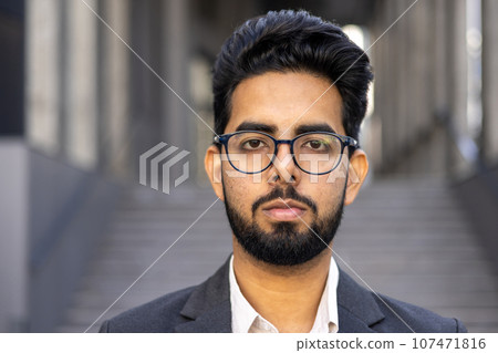 Close up portrait of young successful businessman, thinking serious concentrated man looking at camera outside office building, wearing suit and glasses. Close up portrait of young successful businessman, thinking serious concentrated man looking at camera outside office building, wearing suit and glasses. 107471816
