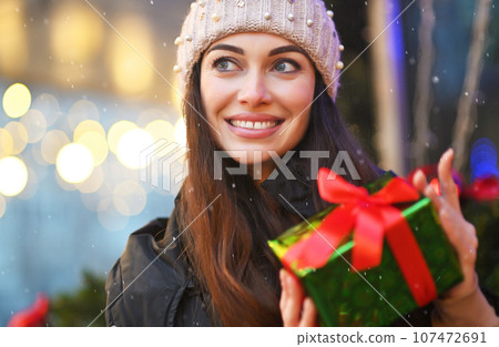 portrait of a young woman in a knitted hat, who smiles and holds a Christmas present in her hands against the backdrop of snow and Christmas lights portrait of a young woman in a knitted hat, who smiles and holds a Christmas present in her hands against the backdrop of snow and Christmas lights 107472691
