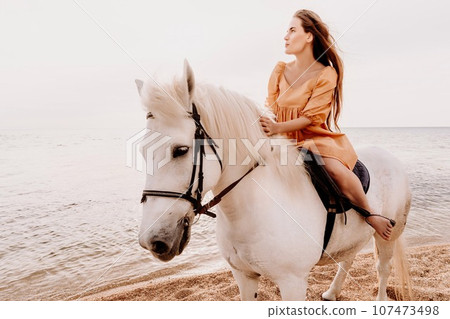 A white horse and a woman in a dress stand on a beach, with the sky and sea creating a picturesque backdrop for the scene. A white horse and a woman in a dress stand on a beach, with the sky and sea creating a picturesque backdrop for the scene. 107473498