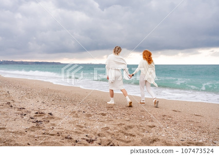Women sea walk friendship spring. Two girlfriends, redhead and blonde, middle-aged walk along the sandy beach of the sea, dressed in white clothes. Against the backdrop of a cloudy sky and the winter Women sea walk friendship spring. Two girlfriends, redhead and blonde, middle-aged walk along the sandy beach of the sea, dressed in white clothes. Against the backdrop of a cloudy sky and the winter 107473524