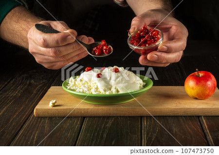 The chef adds jam with a spoon to a dish of cheese and sour cream. The idea of a dietary breakfast on a black kitchen table with the hands of a cook 107473750