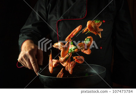 The cook in his hand holds a frying pan with frozen shrimp and parsley. Isolated on black background. Sea food or seafood recipes and restaurant and home cooking 107473784