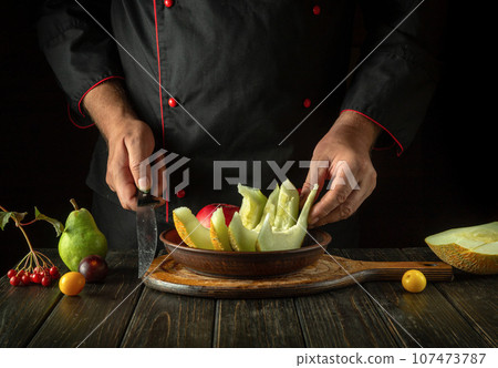 The cook puts fruit on a plate for serving for dessert. Fruit diet concept in hotel kitchen. Asian national cuisine The cook puts fruit on a plate for serving for dessert. Fruit diet concept in hotel kitchen. Asian national cuisine 107473787