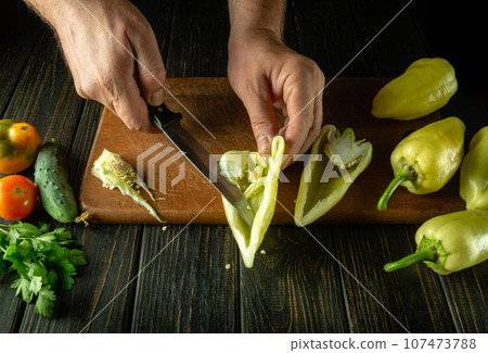 The chef cleans yellow fresh peppers on a cutting board with a knife to prepare a delicious vegetable dish. The concept of cooking vegetable diet on the kitchen table 107473788