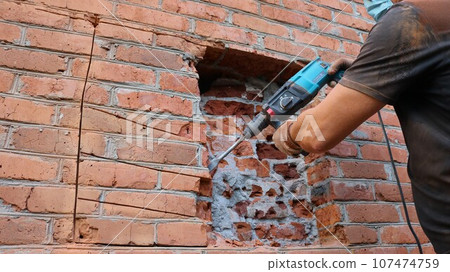 a builder in a black T-shirt and gloves chiseling the brickwork of a house wall with a pneumatic drill, working to destroy a brick wall to make a window opening, drilling the external wall 107474759