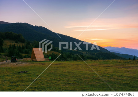 sunset in carpathian mountains. distant rural valley with rolling hills between forested slopes in hazy atmosphere. wooden shelter on the grassy meadow 107474865