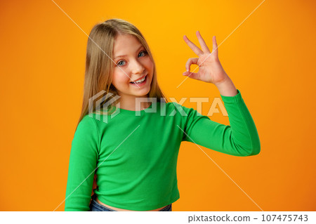 Smiling teen girl showing ok sign against yellow background Smiling teen girl showing ok sign against yellow background 107475743