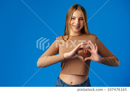 Friendly teen girl shows heart love gesture on blue background Friendly teen girl shows heart love gesture on blue background 107476165