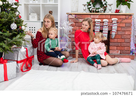 Two women sit together with their young children by the Christmas tree 107476671