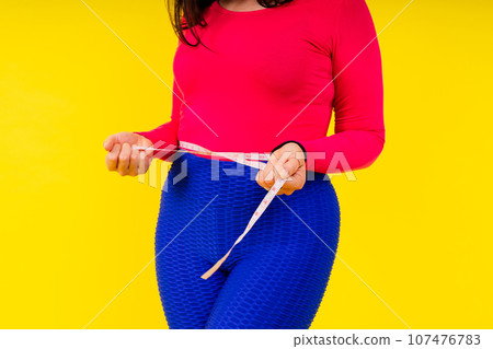Overweight woman measuring waist before weight loss in studio shot on yellow background 107476783