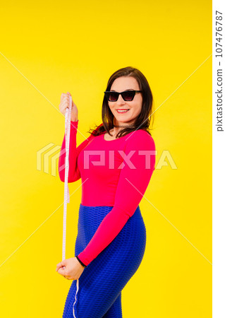 Overweight woman measuring waist before weight loss in studio shot on yellow background 107476787