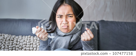Cost of living and heating problems. Close up of angry girl sneezing, staying at home and freezing, wearing warm clothes and scarf Cost of living and heating problems. Close up of angry girl sneezing, staying at home and freezing, wearing warm clothes and scarf 107477158