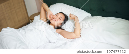 Happy mornings. Beautiful asian girl wakes up in a hotel suit, lying in white bed, stretching her arms and smiling happily 107477312