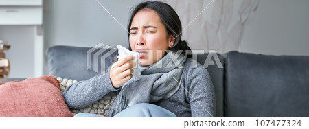 Close up of korean girl sneezing in napking, sitting at home in scarf, feeling unwell, concept of winter, heating cost and people 107477324