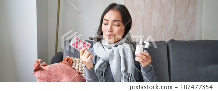 Korean woman showing medical pills, sore throat sprays, feeling sick, staying at home as feeling unwell, catching cold in winter 107477354