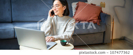Portrait of smiling asian woman making phone call, working on laptop and having conversation on smartphone Portrait of smiling asian woman making phone call, working on laptop and having conversation on smartphone 107477362