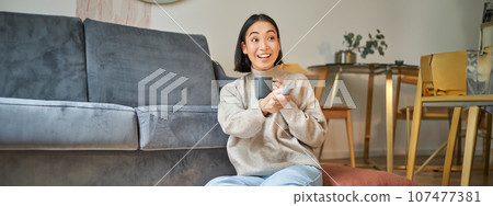 Portrait of smiling young woman resting at home near tv, watching television sitting on floor and drinking coffee from cup 107477381