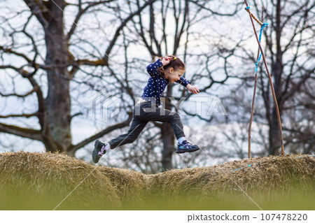 Active little girl jumping between hay with happy facial expression and bright smile, enjoying leisure on autumn day. 107478220