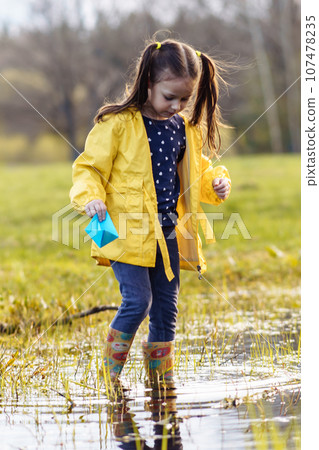 Tranquile child standing in puddle with paper boat in hand and looking at water, preparing to launch ship in fall day. Tranquile child standing in puddle with paper boat in hand and looking at water, preparing to launch ship in fall day. 107478235