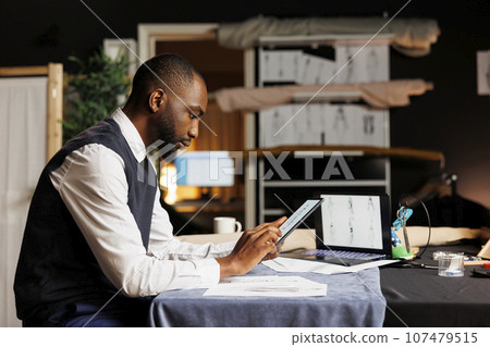 Creative fashion designer in professional tailoring studio meticulously examining handdrawn sketches of bespoken sartorial outfits, seeking inspiration for next collection 107479515