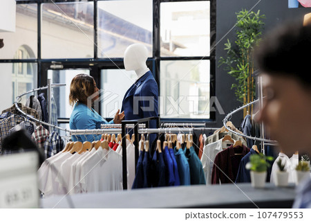 Stylish woman shopping in clothing store, looking at mannequin with formal suit. Shopaholic african american woman analyzing fashionable merchandise fabric in modern boutique. Fashion concept Stylish woman shopping in clothing store, looking at mannequin with formal suit. Shopaholic african american woman analyzing fashionable merchandise fabric in modern boutique. Fashion concept 107479553