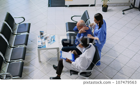 Asian nurse helping senior patient to take off cervical neck collar in hospital reception, african american doctor explaining medical expertise. Elderly man having checkup visit consultation 107479563