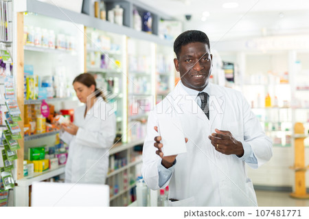 Portrait of african american male pharmacist in pharmacy demonstrating goods 107481771