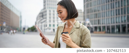 Portrait of smiling korean girl looks at tablet, reads on street, drinks takeaway coffee 107482513