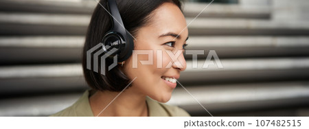Close up portrait of smiling asian girl in headphones, listens to music outdoors, looking happy. 107482515