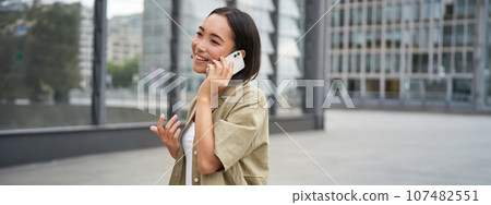Cellular connection. Young asian woman makes a telephone call, talking on mobile smartphone and walking on street 107482551