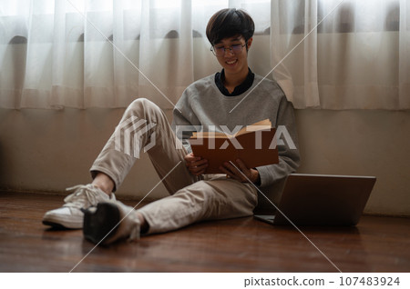 A happy young Asian man enjoys reading a book while sitting on the floor in a room. A happy young Asian man enjoys reading a book while sitting on the floor in a room. 107483924