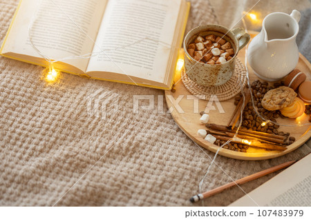 Top view of an opened book, a hot cocoa mug, and accessories are on a bed in a cosy bedroom. 107483979