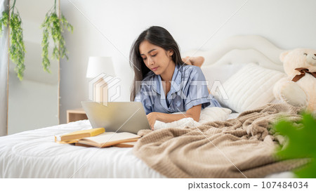 A beautiful Asian woman in pajamas is focusing on her work on her laptop while lying on her bed. 107484034