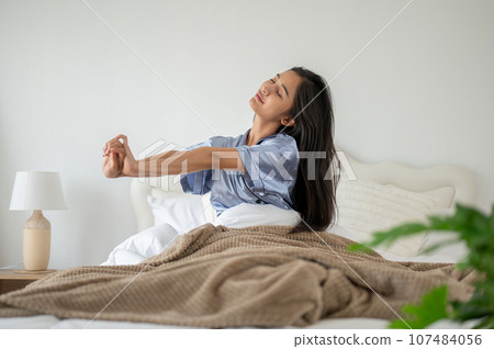 A relaxed and carefree Asian woman in pajamas is stretching her arms after waking up in the morning. 107484056