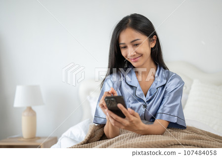 A beautiful Asian woman is using her phone while sitting on her bed after waking up in the morning. 107484058