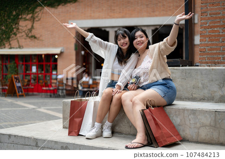 Two attractive Asian female friends are sitting on the stairs in the city with their shopping bags. 107484213
