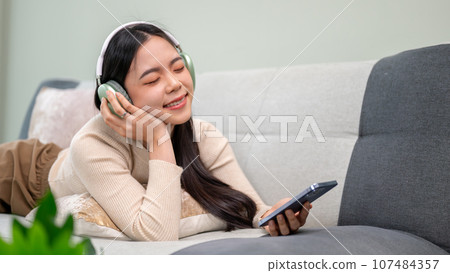 A relaxed and beautiful Asian woman wearing headphones and enjoying the music while lying on a sofa. 107484357