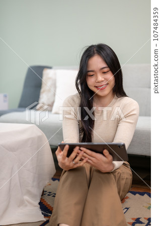 A beautiful Asian woman watching a movie on her digital tablet while relaxing in her living room. 107484359