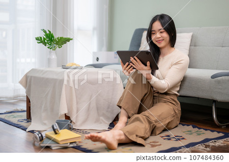 A beautiful Asian woman watching a movie on her digital tablet while relaxing in her living room. 107484360