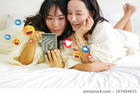 Two cheerful asian women, nestled in bed in bathrobes, are indulged in gleeful selfie-session, surrounded by vibrant digital emoticons. 107484653