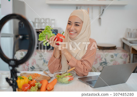 A beautiful Asian Muslim female food blogger is live streaming on her social media in the kitchen. A beautiful Asian Muslim female food blogger is live streaming on her social media in the kitchen. 107485280
