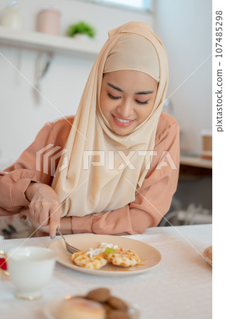 A beautiful Asian Muslim woman eating waffles, having her breakfast at a table in the kitchen 107485298