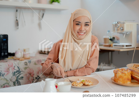 A happy Asian Muslim woman eating waffles at a table in the kitchen. Homemade pastry 107485299