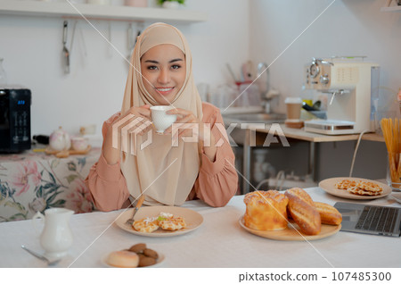 A happy Asian Muslim woman is sipping hot tea and having breakfast at a table in the kitchen. 107485300