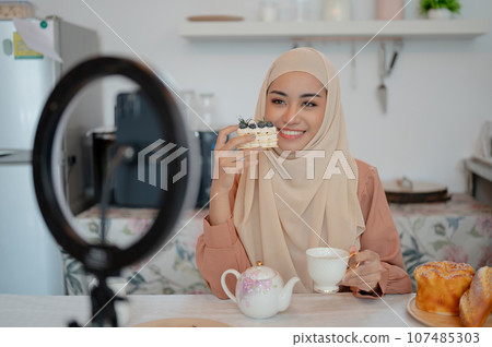 A beautiful Asian Muslim female food blogger is eating a yummy cake and hot tea at a table A beautiful Asian Muslim female food blogger is eating a yummy cake and hot tea at a table 107485303