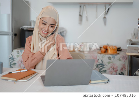 A beautiful Asian Muslim woman is talking on the phone while working on her laptop in the kitchen 107485316